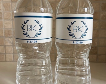 May include: Two clear plastic water bottles with white labels featuring a navy blue wreath design and the initials "BK". The date "9.17.29" is printed below the initials. The bottles are filled with water and have white caps.