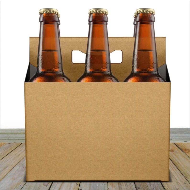May include: A brown cardboard six-pack carrier containing three brown glass beer bottles with gold bottle caps. The bottles are filled with a golden liquid. The carrier is on a wooden surface.