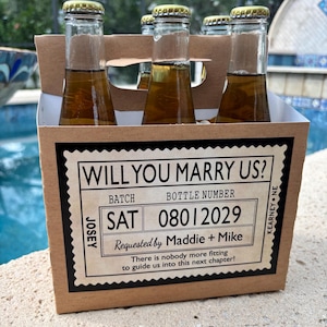 May include: A six-pack of bottled beverages in a cardboard carrier. The carrier has a label that reads "WILL YOU MARRY US?" with the date "08012029" and the names "Maddie + Mike". The bottles have gold caps.