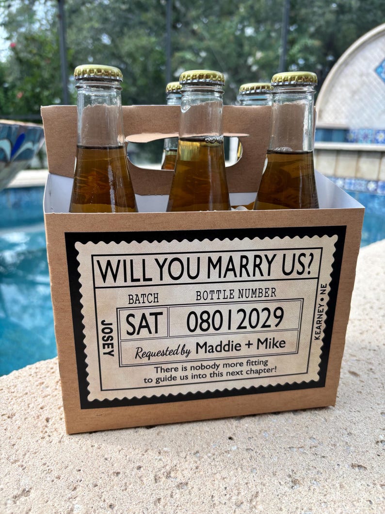 May include: A six-pack of bottled beverages in a cardboard carrier. The carrier has a label that reads "WILL YOU MARRY US?" with the date "08012029" and the names "Maddie + Mike". The bottles have gold caps.