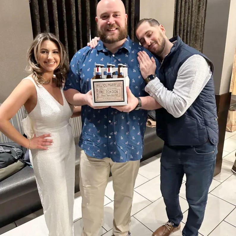 May include: A group of three people celebrating. A man holds a six-pack of beer in a custom box that reads "Will You Marry Us?" and "Fri 7/24/2026". The woman wears a white dress. The man on the right wears a blue vest.