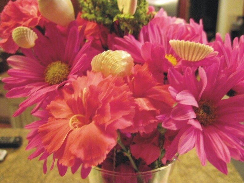 May include: A bouquet of pink and red flowers with seashells and pearls as accents. The flowers are arranged in a clear glass vase.