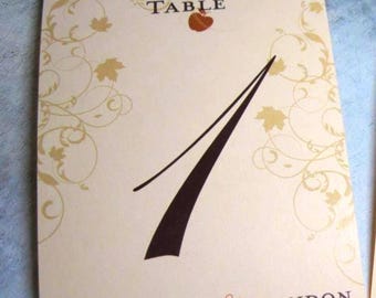May include: A cream-colored table card with the number 1 in dark brown. The word "TABLE" is at the top, with a maple leaf design. The names "Michelle & Brandon" and the date "10.16.11" are at the bottom, with gold and brown accents.