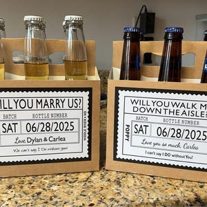 May include: Two cardboard beer carriers with custom labels. One reads "Will You Marry Us?" and the other "Will You Walk Me Down the Aisle?" Each holds beer bottles. The labels include a date, names, and a message.