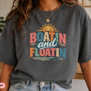 Peut inclure: Un t-shirt gris anthracite à manches courtes avec une impression graphique représentant un soleil souriant au-dessus du texte "BOATIN and FLOATIN" en lettres colorées et usées, avec des vagues stylisées au bas du motif.