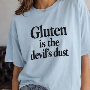 May include: A light blue short-sleeve t-shirt featuring the black printed text "Gluten is the devil's dust." The shirt has a crew neckline and a relaxed fit, constructed from a solid-colored fabric with a smooth texture and visible stitching.