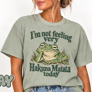 Op de afbeelding: Een saliegroen T-shirt met korte mouwen en een afbeelding van een rustende kikker in het gras. De tekst boven de kikker luidt "I'm not feeling very" en de tekst eronder "Hakuna Matata today" in een donkergroen, verweerd lettertype in vintage-stijl.