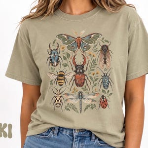 Könnte beinhalten: Ein khakifarbenes Kurzarm-T-Shirt mit einem zentralen Grafikdruck verschiedener Insekten, darunter ein großer Nachtfalter, ein Hirschkäfer, eine Libelle, eine Biene und ein Marienkäfer, umgeben von kleinen botanischen Illustrationen.