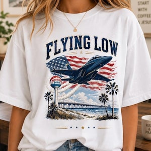 May include: A white t-shirt featuring a graphic print with the text FLYING LOW in dark blue letters. The design includes a dark blue fighter jet flying over a beach scene with a Pensacola Beach water tower, a pier, palm trees, and an American flag.