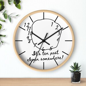May include: A round wall clock with a light wood frame and a white face. The clock features black hour markers and hands. In the center, a hand-drawn circle contains numbers 1 through 12, and the text It's ten past eleven somewhere!