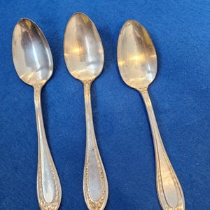 Puede incluir: Tres cucharas de metal plateado dispuestas verticalmente. Cada cuchara presenta un cuenco ovalado liso y un mango delgado decorado con un borde de cuentas y un pequeño motivo floral en relieve en la base del mango.
