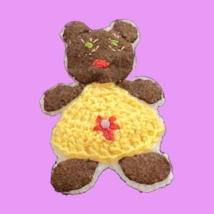 May include: A handmade teddy bear-shaped applique with a brown head and limbs, a yellow crocheted dress, and a red flower. The bear has green stitched eyes and a red mouth. The applique is outlined in white and set against a lavender background.