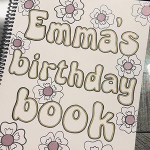 May include: A white book with a gold outline and the text "Emma's birthday book" in a whimsical font. The book is decorated with white flowers with pink centers.