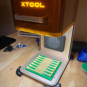 Pode incluir: Uma máquina de gravação a laser com a palavra "XTOOL" iluminada em laranja. A máquina está aberta, revelando uma bandeja verde com vários tees de golfe de madeira. A máquina está sobre uma superfície de madeira.