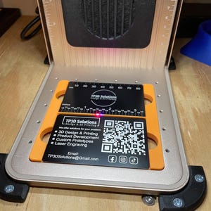 Pode incluir: Uma máquina de gravação a laser com um porta-cartões de visita preto e laranja. O cartão de visita tem o texto "TP3D Solutions" e um código QR. A máquina tem uma régua com medidas em polegadas.