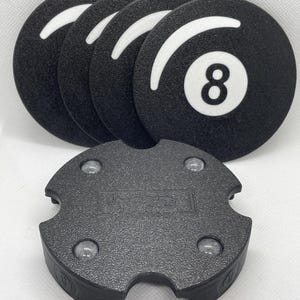 May include: A set of four black round coasters with a white 8-ball design, and a black, textured, four-pronged base. The coasters have a white curved stripe. The base has the word "TETON" printed on it.