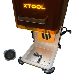 Puede incluir: Una máquina de grabado láser con la palabra "XTOOL" iluminada en amarillo. La máquina tiene una parte superior marrón y una base plateada con un pequeño indicador digital de temperatura y humedad. La máquina tiene una ventilación y un asa.