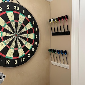 Może przedstawiać: Tarcza do darta z czarnym, czerwonym, zielonym i żółtym wzorem jest zamontowana na jasnobrązowej ścianie. Dwa uchwyty na rzutki z rzutkami są zamontowane na ścianie. Rzutki mają lotki w różnych kolorach.