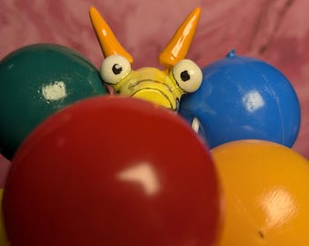 May include: A whimsical toy with a yellow body, orange horns, and large eyes, holding onto a cluster of colorful balloons. The balloons are red, blue, green, and orange.
