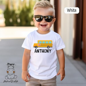 May include: White short-sleeved t-shirt with a yellow school bus graphic and the name "ANTHONY" printed in black.