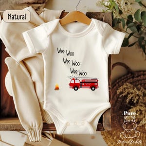 May include: A white baby onesie with short sleeves features a red fire truck with a ladder and the words "Wee Woo Wee Woo Wee Woo" printed on the front.