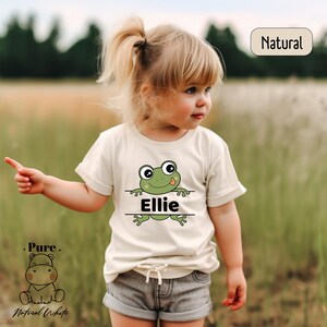 a little girl standing in a field with a frog on her shirt