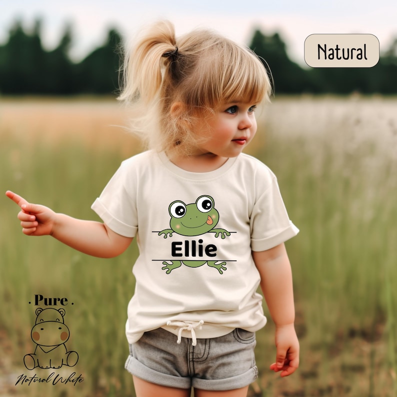 a little girl standing in a field with a frog on her shirt