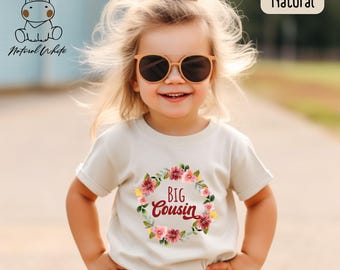 Könnte beinhalten: Ein kleines Kind trägt ein weißes T-Shirt mit einem Blumengirlanden-Design und dem Text "BIG COUSIN" in Rot. Das Kind trägt eine Sonnenbrille und hat die Haare zu einem Pferdeschwanz gebunden.