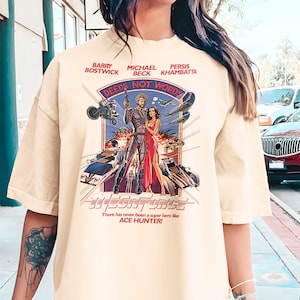 Peut inclure: T-shirt crème avec un design de poster de film rétro. Le graphisme comprend le texte "DEEDS NOT WORDS" et "MEGAFORCE", avec des images de personnages et de véhicules. Le t-shirt contient également le texte "There has never been a super hero like ACE HUNTER!"