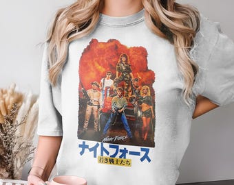 Puede incluir: Camiseta gris claro con un diseño de cartel de película que presenta a un grupo de personas frente a una explosión naranja. El texto japonés dice "Night Force" y "Jóvenes guerreros."