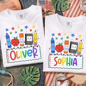 May include: Two white t-shirts featuring a colorful school supply graphic with a blue crayon, red apple, orange glue stick, black and white composition notebook, and yellow pencil. The names Oliver and Sophia are printed in rainbow letters.