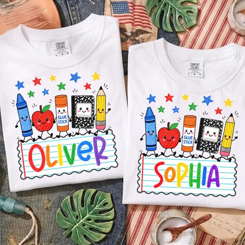 May include: Two white t-shirts featuring a colorful school supply graphic with a blue crayon, red apple, orange glue stick, black and white composition notebook, and yellow pencil. The names Oliver and Sophia are printed in rainbow letters.