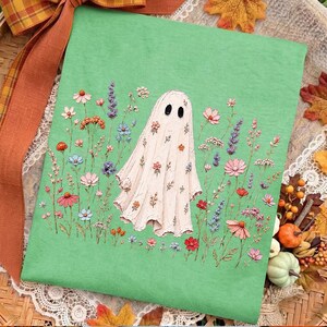 May include: A light green t-shirt featuring a central graphic of a white ghost covered in small colorful flowers. The ghost is surrounded by a field of various wildflowers in shades of pink, blue, orange, and purple, creating a detailed botanical scene.