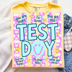May include: Yellow t-shirt with the words "TEST DAY" in large, light blue letters. The shirt has a floral and striped design with positive affirmations like "You are smart!" and "You got this!"