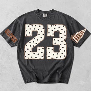 May include: A charcoal gray short-sleeve t-shirt featuring the large white number 23 with black polka dots in the center. The right sleeve displays the name OLIVER in brown, and the left sleeve features a brown pennant graphic with the word EAGLES.