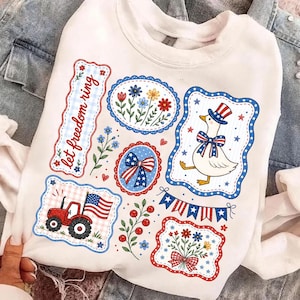 May include: White sweatshirt with patriotic themed graphics. Features a red tractor with an American flag, a goose in a top hat, floral designs, and the text "let freedom ring". The graphics are outlined in red and blue.