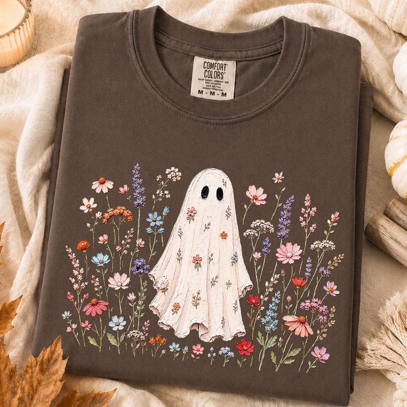 May include: A dark brown crewneck t-shirt featuring a central graphic of a white ghost surrounded by a colorful array of wildflowers in shades of pink, blue, orange, and purple. The interior neck label displays the text Comfort Colors size M.
