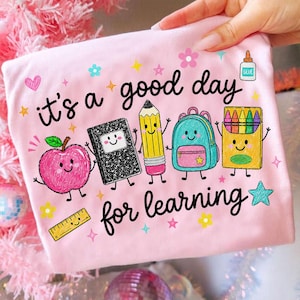 May include: A light pink t-shirt featuring the black text "it's a good day for learning" and colorful cartoon illustrations of a smiling apple, a composition notebook, a pencil, a backpack, a crayon box, a glue bottle, and a ruler.