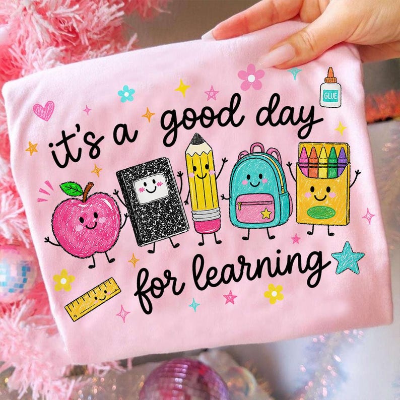 May include: A light pink t-shirt featuring the black text "it's a good day for learning" and colorful cartoon illustrations of a smiling apple, a composition notebook, a pencil, a backpack, a crayon box, a glue bottle, and a ruler.