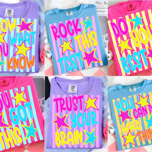 May include: A collection of six short-sleeve t-shirts in purple, blue, and pink, each featuring vertical stripes and colorful star graphics. The shirts display motivational phrases including Show What You Know, Rock This Test, Do Your Best, You Got This, Trust Your Brain, and You Can Do Hard Things.