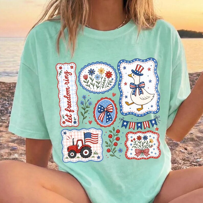 May include: A light green t-shirt featuring patriotic designs. The shirt has various framed illustrations, including a goose in a top hat, a tractor with an American flag, and floral arrangements. The text "let freedom ring" is also displayed.