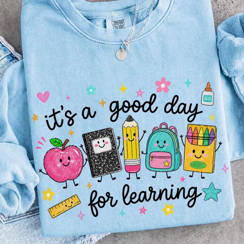 May include: A light blue crewneck sweatshirt featuring a graphic print of a smiling apple, notebook, pencil, backpack, crayon box, and ruler. The text reads, "it's a good day for learning." Small colorful stars and flowers surround the school supplies.