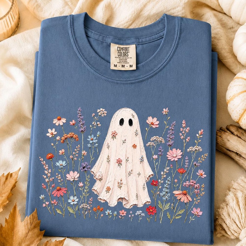 May include: A blue crew neck t-shirt with a Comfort Colors label at the inner collar. The front features a graphic of a white ghost surrounded by a variety of colorful wildflowers in shades of pink, orange, blue, and purple, with small floral patterns on the ghost.