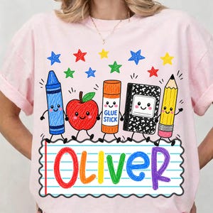 May include: A light pink t-shirt featuring a colorful graphic of a blue crayon, a red apple, an orange glue stick, a black and white composition notebook, and a yellow pencil, all with faces, above the name Oliver written in rainbow letters.