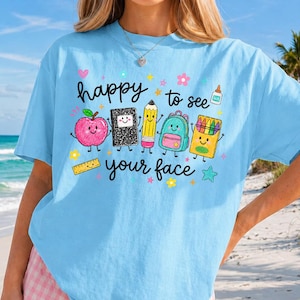 Puede incluir: Una camiseta azul claro con un estampado gr&aacute;fico de &uacute;tiles escolares con caras, incluyendo una manzana, un cuaderno, un l&aacute;piz, una mochila y ceras. El texto "happy to see your face" aparece en negro, rodeado de estrellas y flores de colores.