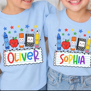 May include: Two light blue t-shirts featuring a colorful school-themed graphic. The design includes a blue crayon, red apple, orange glue stick, black composition notebook, and yellow pencil with faces, above a lined paper banner with names.