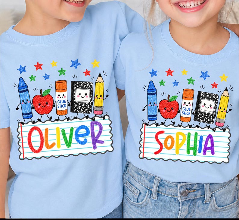 May include: Two light blue t-shirts featuring a colorful school-themed graphic. The design includes a blue crayon, red apple, orange glue stick, black composition notebook, and yellow pencil with faces, above a lined paper banner with names.