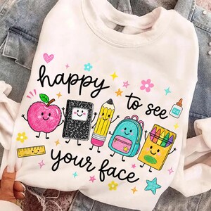 Puede incluir: Una sudadera blanca de manga larga con un gr&aacute;fico colorido de &uacute;tiles escolares con caras, incluyendo una manzana, un cuaderno, un l&aacute;piz, una mochila y crayones. El texto "happy to see your face" aparece en letra cursiva de color negro.