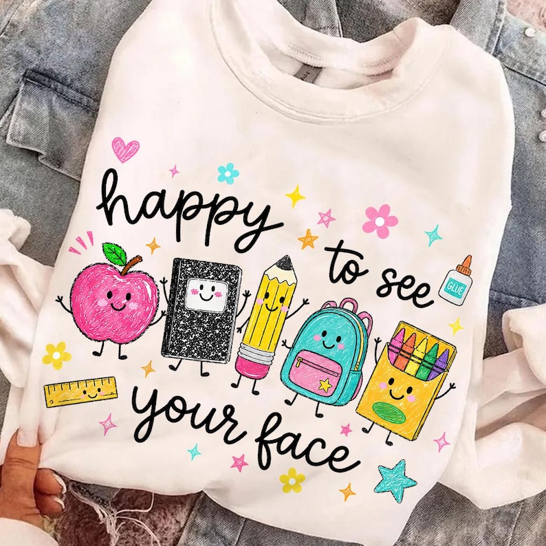 Puede incluir: Una sudadera blanca de manga larga con un gr&aacute;fico colorido de &uacute;tiles escolares con caras, incluyendo una manzana, un cuaderno, un l&aacute;piz, una mochila y crayones. El texto "happy to see your face" aparece en letra cursiva de color negro.