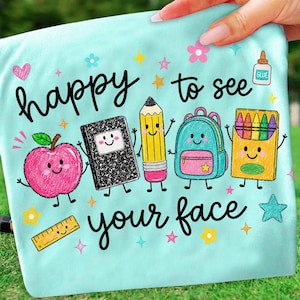 Puede incluir: Una camiseta azul claro con un gr&aacute;fico colorido de &uacute;tiles escolares con caras, incluyendo una manzana, un cuaderno, un l&aacute;piz, una mochila y crayones. El texto "happy to see your face" aparece en letra cursiva negra, rodeado de estrellas y flores.